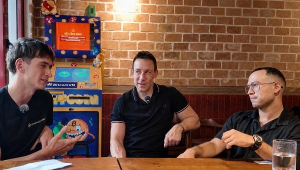Three people sitting at a wooden table beside a BitcoinVN BTM in a brick-walled cafe setting.