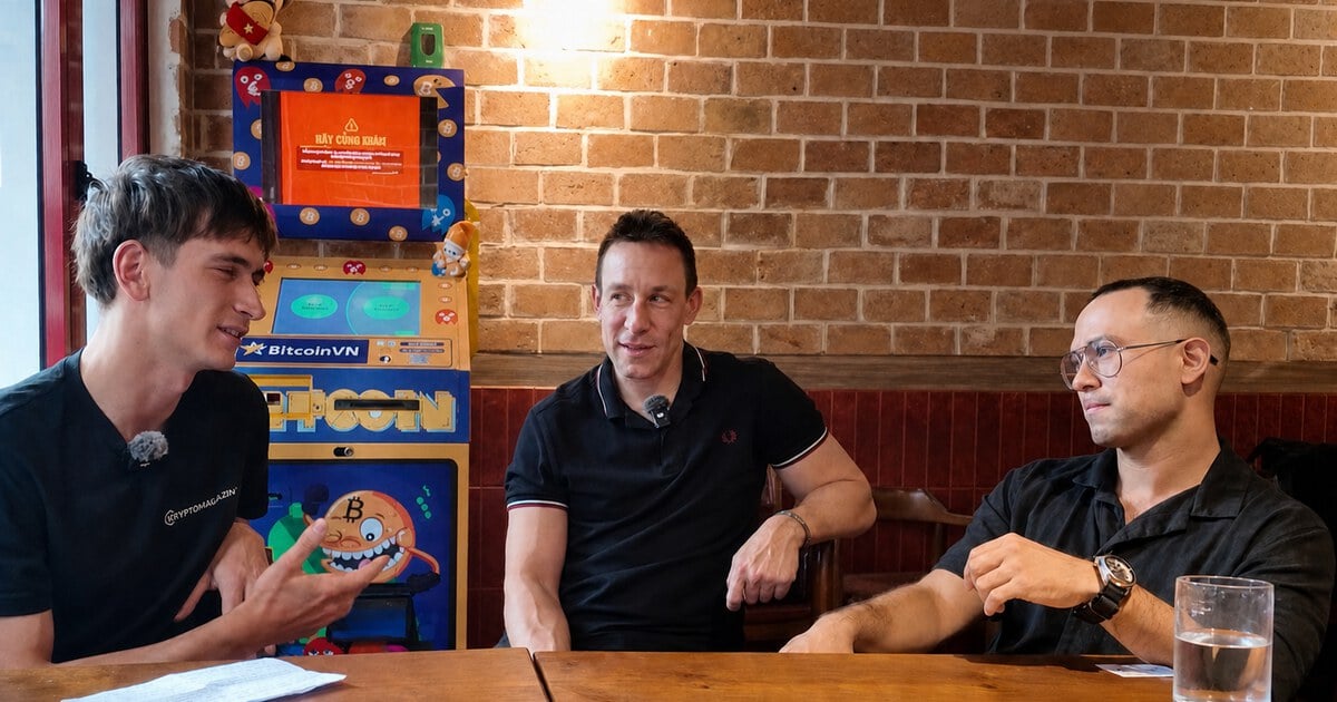 Three people sitting at a wooden table beside a BitcoinVN BTM in a brick-walled cafe setting.